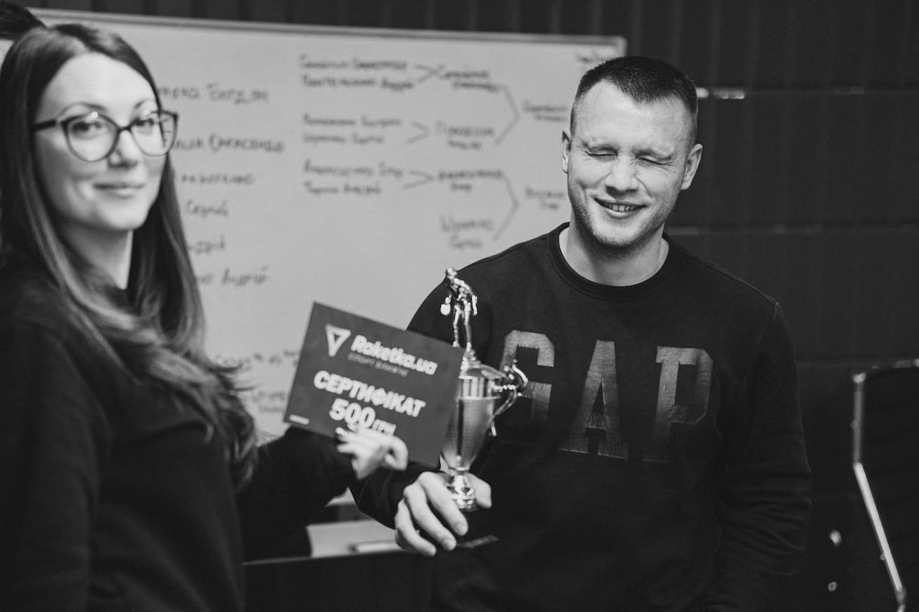 Serhii Shramko smiling and holding a trophy, standing next to a woman holding a 500 UAH certificate in a casual office setting with a whiteboard in the background.